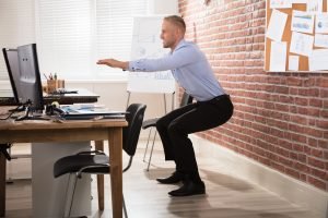 Office worker standing and stretching beside a desk, demonstrating movement and mobility exercises to counteract long hours of sitting.