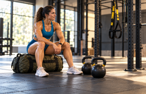 Female athlete resting after training in a gym, seated on a sandbag beside two kettlebells, with suspension straps hanging nearby.