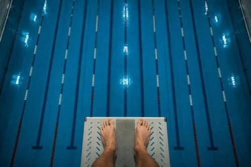 A diver stands on the edge of a high diving board looking down into the water of an Olympic-sized swimming pool.