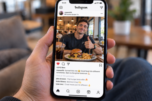 A hand holds a smartphone displaying an Instagram post of a smiling male athlete in a burger restaurant giving a thumbs-up beside a large burger, fries and a soft drink, illustrating the idea of enjoying an indulgent meal within a balanced diet.