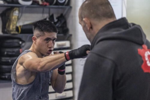 Young boxer punching heavy bag with coach holding it, symbolising training and discipline in Western Boxing.
