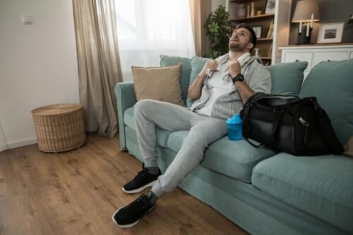 Man relaxing on couch next to gym bag procrastinating instead of training showing comfort zone behaviour