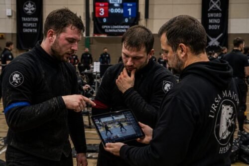 HEMA practitioners review footage of a recent tournament bout on a tablet, discussing techniques, timing, positioning, and tactical decisions while analysing the exchange.