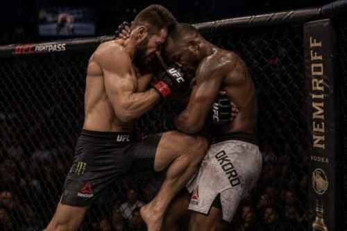 MMA fighter controls an opponent against the cage with head position and an underhook while driving a knee.