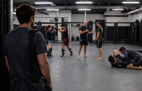Newcomer carrying a gym bag watches a structured MMA class with pad work, coaching and controlled grappling.