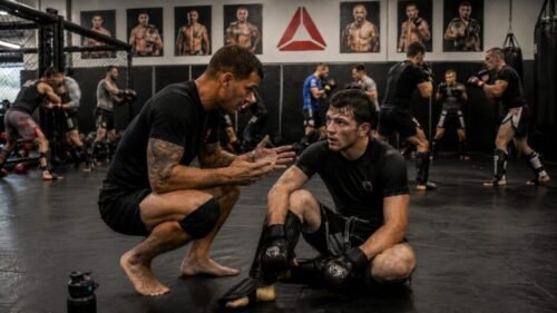 Tired MMA beginner sits on the mat while a coach crouches beside him and explains a technical correction.