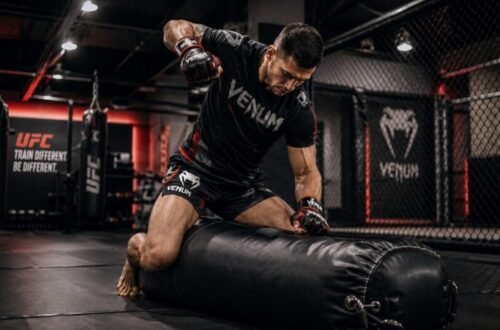 MMA trainee wearing branded black and red equipment straddles a heavy bag on the floor and practises ground-and-pound.