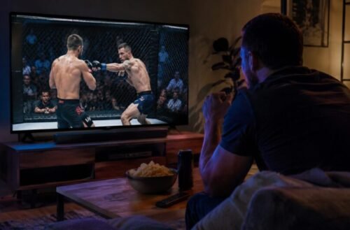 MMA fan leans towards a television while watching two fighters exchange strikes inside the cage during a live event.