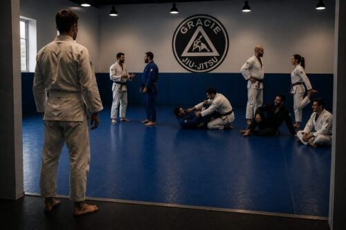 New white belt standing at the edge of a Brazilian Jiu-Jitsu academy, watching students prepare for class while an instructor speaks with practitioners on the mats.