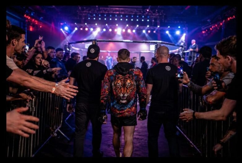 Rear view of an amateur MMA fighter in a colourful walkout jacket approaching the cage with his coaches as spectators lean over barriers under bright arena lights.