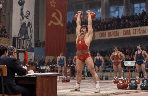 Soviet kettlebell athlete lifting two competition kettlebells overhead during an organised competition in a 1980s sports hall.