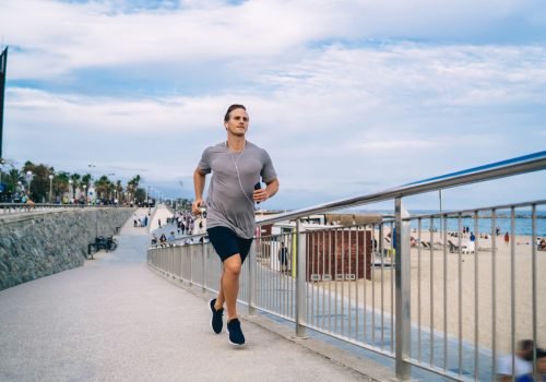 Caucasian bodybuilder running at embankment enjoying leisure training for getting power energy, male athlete in electronic headphones jogging during morning workout keeping wellness and vitality
